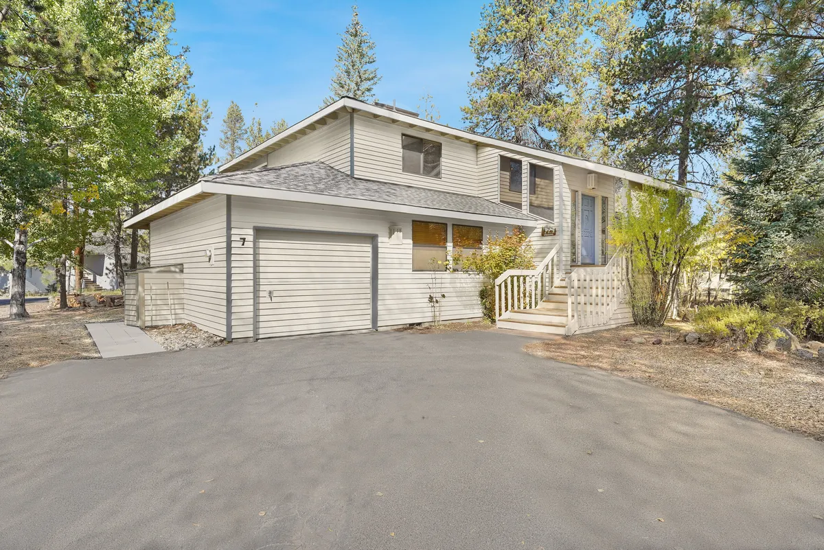 This image features the welcoming front porch of Rogue 7, with inviting landscaping and large windows that hint at the home's connection to nature. The front area is framed by mature trees and greenery, suggesting easy access to outdoor spaces and trails.