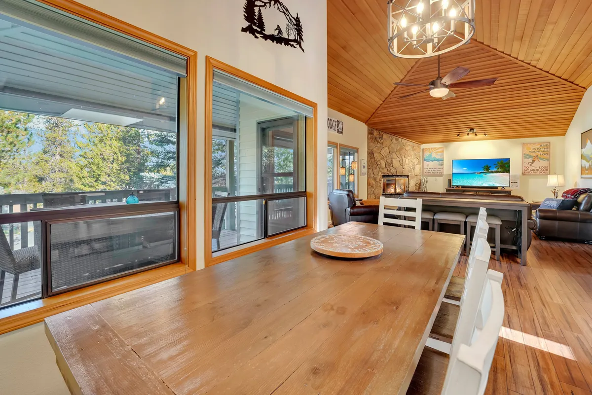 An inviting dining area featuring a large wooden table surrounded by chairs, set beneath a beautiful chandelier. Floor-to-ceiling windows frame scenic views and let in natural light, while a stone accent wall and adjacent TV area add a sense of comfort and sophistication. The setup is perfect for group meals or game nights.