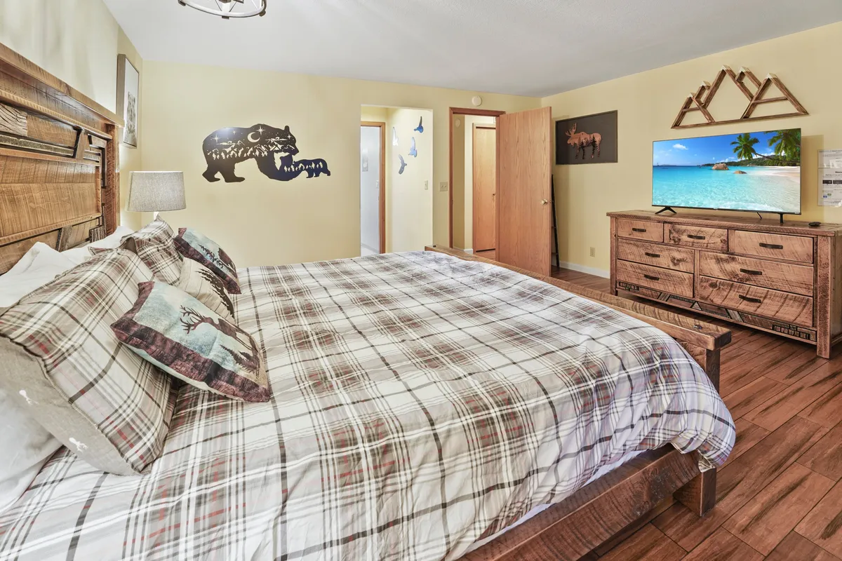 This image features a large bed with plaid bedding in a warmly decorated bedroom. A wooden dresser and decorative animal-themed wall art create a cozy, inviting atmosphere, while a television is positioned for comfortable viewing. Natural light fills the room, enhancing the sense of relaxation and comfort.